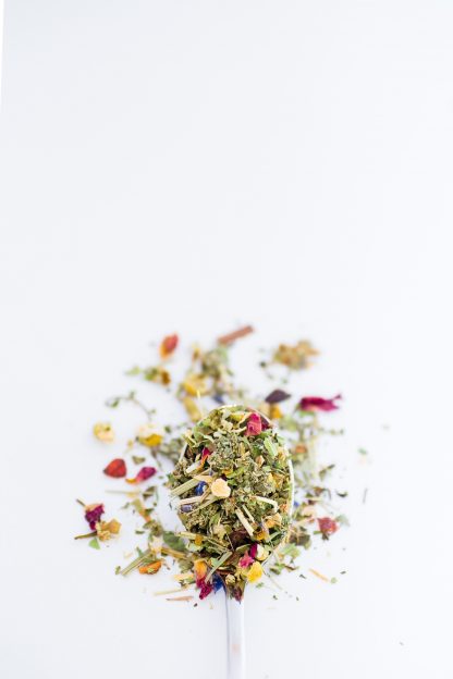 Beautiful dried chamomile florets blended with rose petals, lemon peal, lemon verbena, lemon grass overflow a silver spoon onto a white background