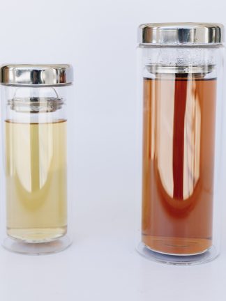 One 12 ounce and one 16 ounce clear glass double walled tea tumbler containing steeped green tea and steeped black tea with a stainless steep strainer separating the liquid and the stainless steel lid on a white background