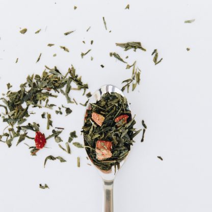 Green tea and strawberry pieces overflow the silver spoon onto a white background